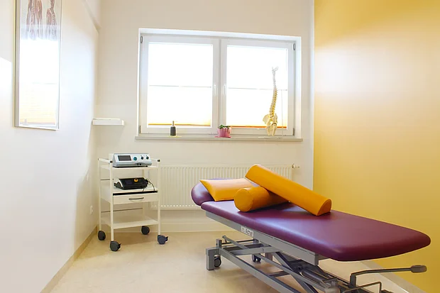 Heller Behandlungsraum mit lila Therapieliege, gelben Lagerungsrollen und Elektrotherapiegerät