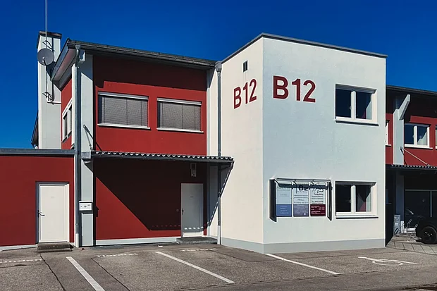 Therapiezentrum B12 von der Straße aus: weißer Gebäudeteil mit Schriftzug B12, davor Parkplätze direkt am Haus, darunter ein Behindertenparkplatz