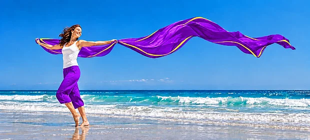 Lächelnde Frau läuft barfuß am Strand und hält ein langes lilafarbenes Tuch, das hinter ihr im Wind weht
